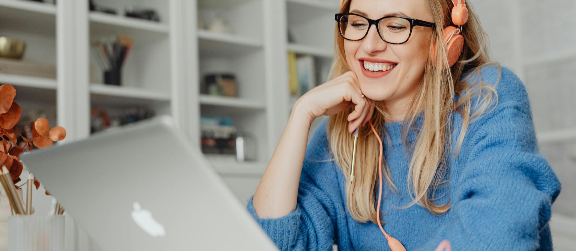 Smiling woman wearing headphones and glasses, working on a laptop in a bright indoor setting.