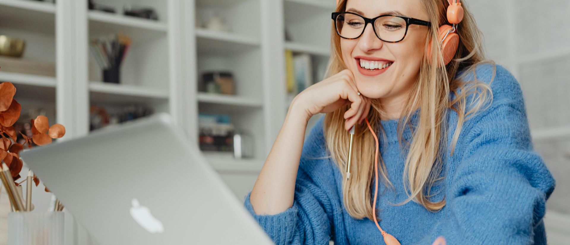 Smiling woman wearing headphones and glasses, working on a laptop in a bright indoor setting.